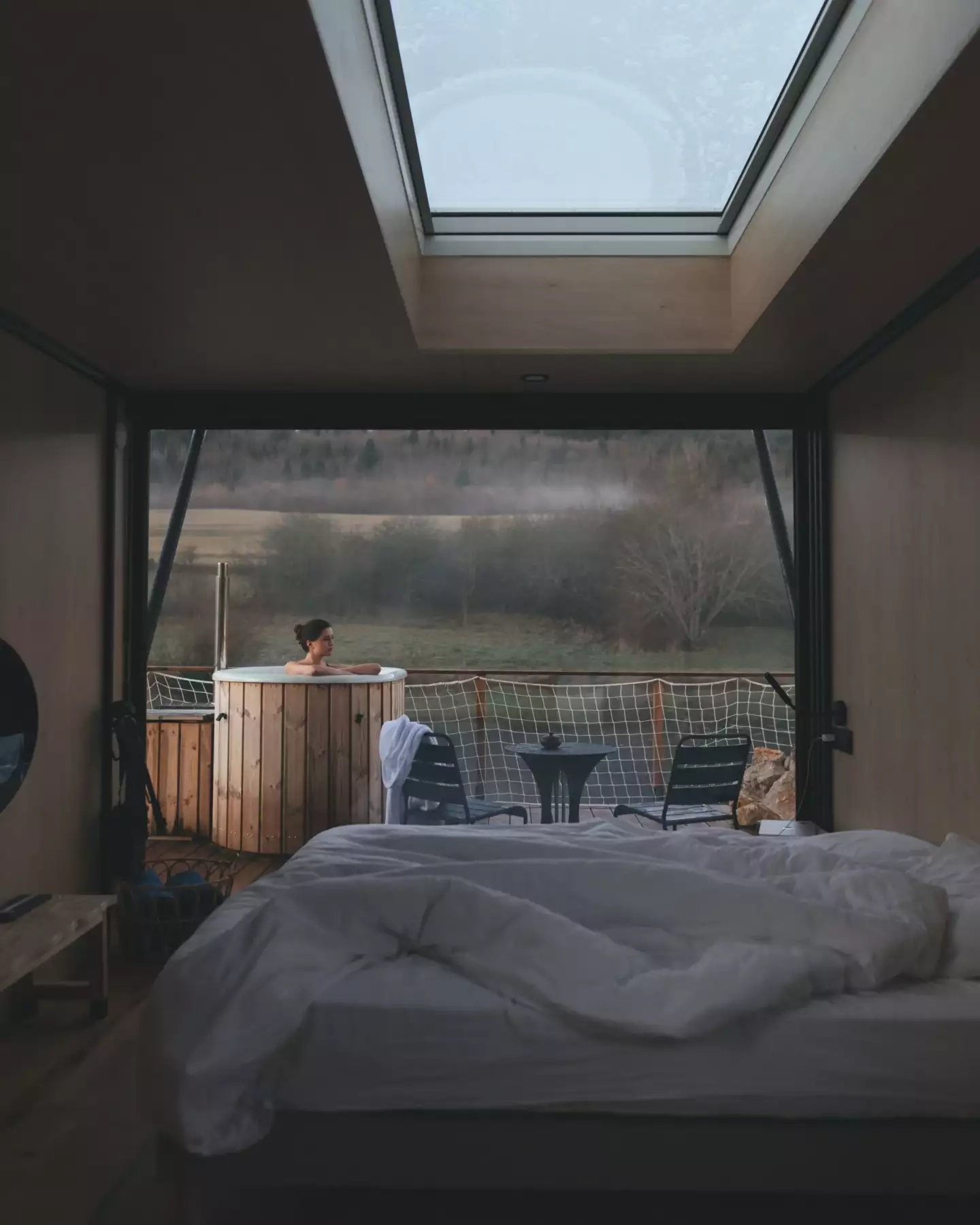 Photo de l'intérieur d'un lodge donnant sur la terrasse, la lumière est douce, on y aperçoit une femme profitant du bain nordique.